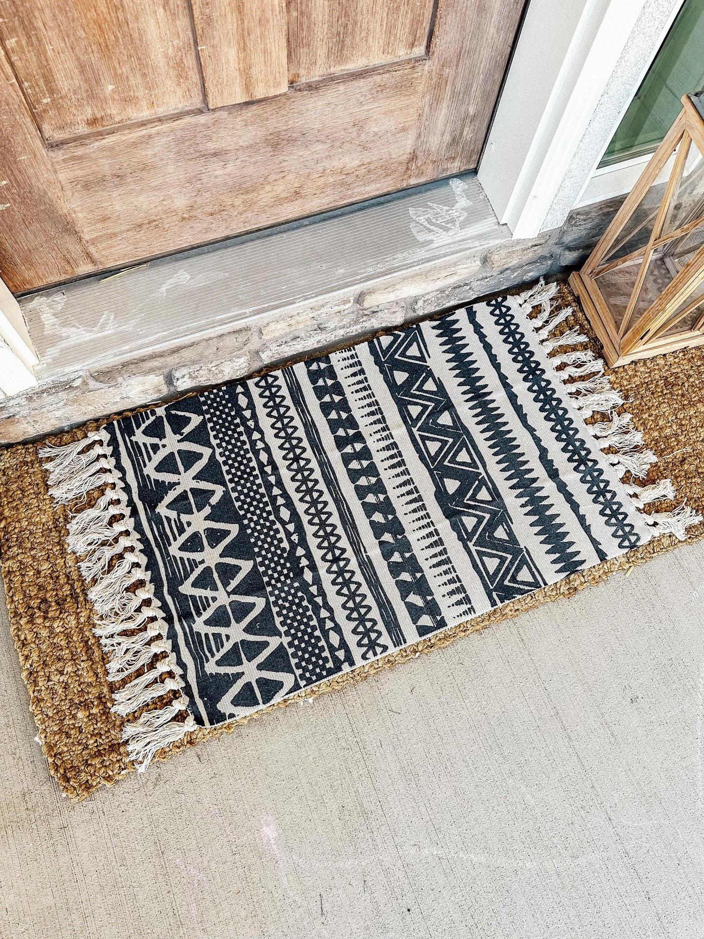 The rug features a black and cream geometric design with various shapes like triangles, zigzags, and lines. It has fringe details on the shorter sides